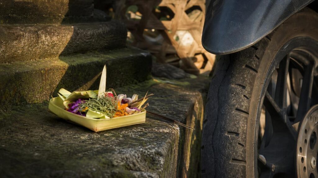 A close textural shot of a Balinese offering basket (canang sari) on a stone step beside a scooter wheel, morning light