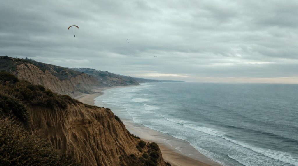 A dramatic coastal cliff overlooking the Pacific Ocean under a soft overcast sky, paragliders as small distant silhouettes