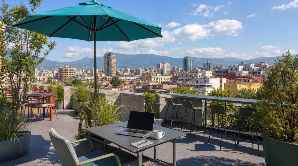 A rooftop coworking terrace in central Tirana at midday, a laptop and coffee cup on a small table, a teal umbrella for shade