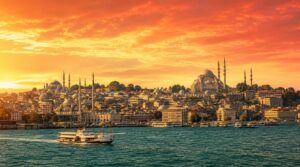 Wide golden-hour skyline shot of Istanbul across the Bosphorus, minarets and mosque domes silhouetted against a warm coral