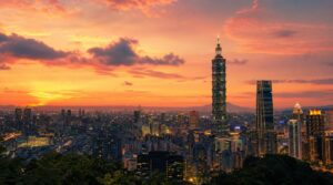 Wide sunset skyline shot of Taipei with Taipei 101 silhouetted against a warm coral-and-golden sky, soft clouds catching the light